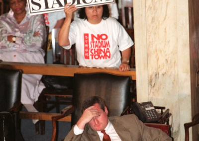 Photo from 2000 of an Asian woman holding a sign that says "chinatown is troubeltown. No Stadium!" at a city council meeting